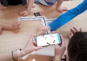 Nauczycielka za pomocą telefonu uruchamia Ozobota. Obok stoją Staś i Hubert , którzy obserwują.