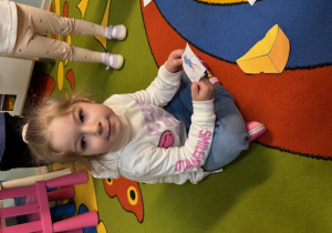 Kornelia siedzi na dywanie, a w ręku ma ilustrację myszy. Obok niej leży ilustracja z kawałkiem sera.