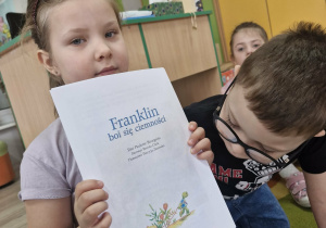 Maja trzyma w dłoniach opowiadania „Franklin boi się ciemności, Antoś ogląda obrazki na okładce opowiadania.