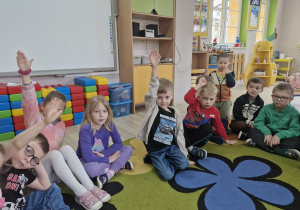 Dzieci siedzą na dywanie trzymając ręce w górze.