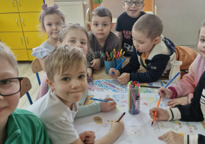 Dzieci siedzą przy stoliku i rysują na dużym kartonie.