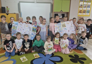 Dzieci stoją przed tablicą multimedialną. Przedszkolaki trzymają w dłoniach wykonane przez siebie dwa duże plakaty.