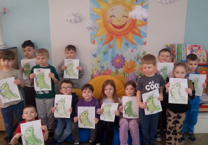 Dzieci prezentują wykonane przez siebie obrazki przedstawiające postać Dinka - maskotki Programu przedszkolnej edukacji antytytoniowej "Czyste powietrze wokół nas". W tle kącik książki, plakat przedstawiający słońce, kwiaty.