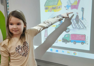 Weronika stoi przed tablicą multimedialną, na której wyświetlony jest plakat "Co dymi?". Dziewczynka wskazuje osoby palące papierosy.