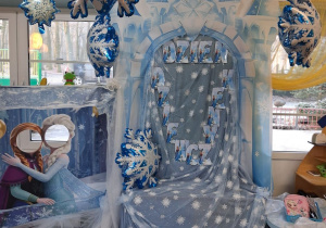 Dekoracja "Kraina lodu" z okazji Dnia Dziewczynek : na tablicy okrytej błekitnym materiałem w białe śnieżynki przypięty jest napis "Dzień Dziewczynek" z motywem "Krainy lodu". Nad tablicą wisi błękitno-biały baner zamku. Z boku stoi fotobudka z motywem "Krainy lodu". Nad dekoracją wiszą balony w kształcie śnieżynek.