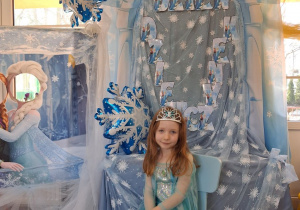 Emma w stroju Elzy pozuje do zdjęcia na tle dekoracji siedząc na błękitnym krzesełku.