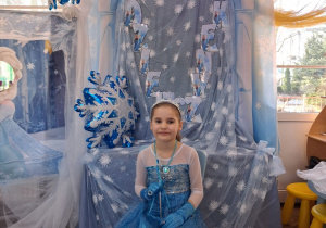 Marysia w stroju Elzy pozuje do zdjęcia na tle dekoracji siedząc na błękitnym krzesełku.