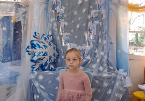 Zuzia pozuje do zdjęcia na tle dekoracji siedząc na błękitnym krzesełku.