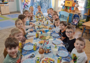 Dzieci siedzą przy wspólnym stole podczas słodkiego poczęstunku. W tle dekoracja z okazji "Dnia Dziewczynek".