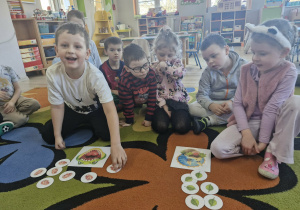 Franuś i Ola siedzą na dywanie. Przed nimi leżą obrazki z wizerunkami dinozaurów oraz kartoniki do zabawy "Karmimy dinozaury".