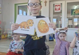 Antek trzyma w dłoniach obrazki z jajem dinozaura i dinozaurem.