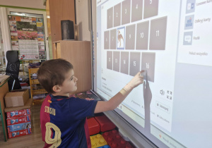 Staś stoi przy tablicy multimedialnej. Chłopiec rozwiązuje dinozaurowe memory.