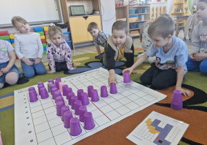 Przedszkolaki układają wizerunek dinozaura na macie do kodowania z wykorzystaniem kolorowych kubeczków.