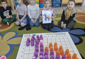 Dzieci siedzą na dywanie. Przed nimi leży rozłożona mata do kodowania, a na niej ułożony z kolorowych kubeczków wizerunek dinozaura.