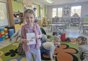 Ola trzyma w dłoniach kartonik z zadaniem dla dzieci. W tle widać przedszkolaki chodzące na czworakach.