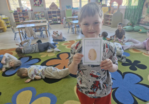 Wojtuś trzyma w dłoniach obrazek z wizerunkiem jaja dinozaura. Za chłopcem widać dzieci turlające się po dywanie jak jajka.