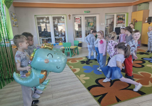 Przedszkolaki stoją na dywanie podczas zabawy ruchowej. Przed nimi Olek i Weronika stoją trzymając balony z dinozaurami.