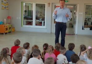 Dzieci siedzą na dywanie, a przed nimi stoi Pan zapraszając przedszkolaków na kolejny występ.