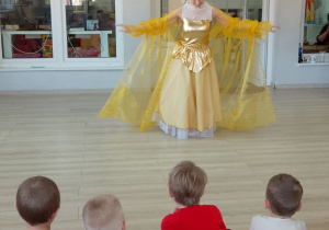 Dzieci siedzą na dywanie, a baletnica w żółtej sukni kłania się po występie.