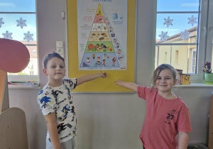 Franek i Weronika wskazują na obrazek Piramidy Zdrowego Żywienia zawieszony na tablicy w sali.