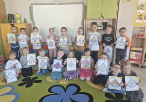 Dzieci stoją przed tablicą multimedialną i prezentują wykonane przez siebie prace plastyczne.