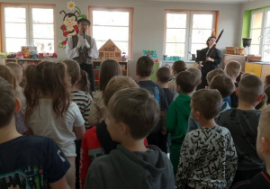 Dzieci śpiewają piosenkę powitalną, a przed nimi stoi Pan prowadzący audycję muzyczną oraz Pani z fagotem. W tle kąciki zabaw.