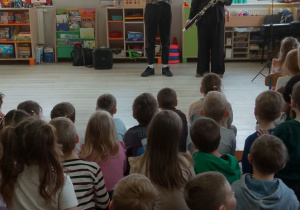 Dzieci siedzą na dywanie, a przed nimi Pani prezentuje instrument dęty - fagot. Obok stoi Pan prowadzący audycję muzyczną.