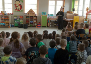 Dzieci siedzą na dywanie, a na środku sceny stoi Pani, która gra na fagocie.