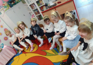 Dziewczynki w odświętnych strojach siedzą na krzesłach z nogą na nodze, na głowach mają diademy w kolorze złota.