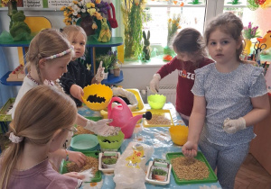 Grupka dziewczynek stoi przy stoliku i nabiera ziemię oraz owies. W tle kącik wielkanocny.