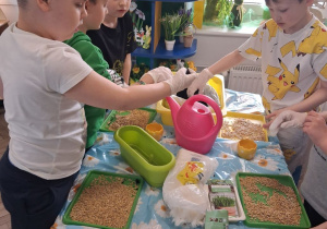 Grupka chłopców stoi przy stoliku i nabiera ziemię. W tle kącik wielkanocny.