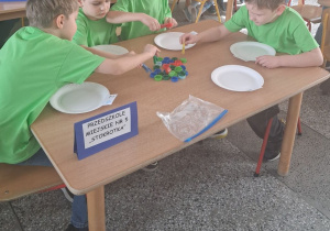 Czterech chłopców w zielonych koszulkach siedzi przy stoliku i za pomocą klamerek do bielizny nakładają kolorowe nakrętki na papierowe talerzyki.