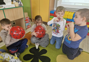 Dzieci siedzą na dywanie. W dłoniach trzymają czerwone balony w czarne kropki, które próbują nadmuchać.