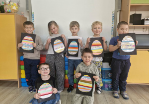 Dzieci stoją przed tablicą multimedialną i prezentują wykonane przez siebie prace plastyczne.