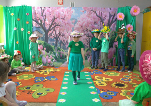 Zuzia idzie po zielonym wybiegu z kwiatkami prezentując swój kapelusz.