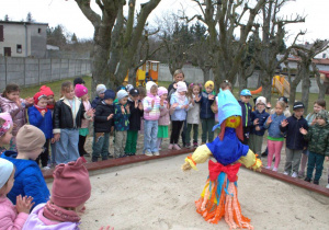 Wszystkie dzieci machają w stronę Marzanny, która znajduje się w piaskownicy na placu zabaw.