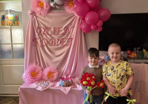 Zdjęcie przedstawia dekoracje w kolorach różu, na różowym tle różowy napis "Dzień kobiet" balony i kwiaty papierowe. Krzyś z Karolem trzymają upominki dla dziewczynek.