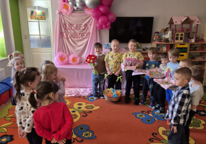 Chłopcy stoją po prawej stronie trzymając upominki dla koleżanek z grupy. Naprzeciwko nich dziewczynki, które uważnie słuchają składanych im życzeń. W tle dekoracja.