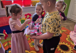 Kornel stoi przed Felicją z tacą, na której znajdują się torebeczki ze słodkościami. Felicja sięga po jedną z nich. Obok niej stoi Wiktoria, która trzyma już swoje słodkości.