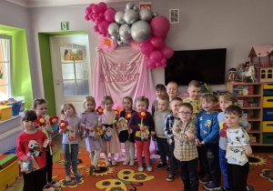 Dziewczynki i chłopcy z grupy stoją na tle dekoracji. Dziewczynki w rękach trzymają prezenty.