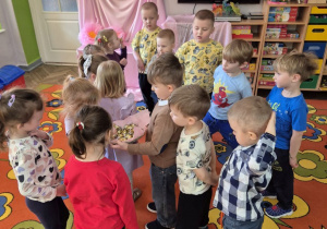 12.Dzieci stoją na dywanie, Krzysio trzyma w rękach koszyk z cukierkami, którymi częstuje koleżanki i kolegów z grupy.