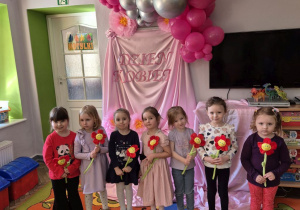 Dziewczynki z grupy stoją na tle dekoracji, w rękach trzymają otrzymane wcześniej pluszowe kwiaty.