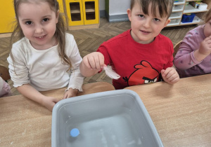 Wiktoria i Krzyś siedzą przy stoliku, przed nimi stoi miska z wodą, na której unosi się plastikowa nakrętka. Krzyś trzyma w ręku białe piórko.