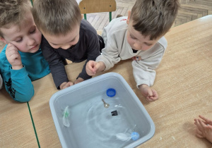 Olek, Filip i Wojtek siedzą przy stoliku i pochylają się nad miską wodą, w której widać wrzucone wcześniej przedmioty.