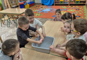 "Motylki" siedzą przy stolikach, a Filip stoi trzymając w dłoniach różową gąbkę, którą wyciska z wody nad miską. Obok niego stoi Wojtek, który już wyciąga rękę po gąbkę.