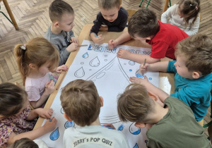 Dzieci stoją dookoła stolika i kolorują niebieskimi kredkami dużą kolorowankę - krople wody.