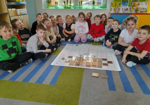 Dzieci siedzą na dywanie, a przed nimi leży mata do kodowania, na której znajduje się ułożony obrazek garnka z brązowych kwadratów i symbole zjawisk atmosferycznych.