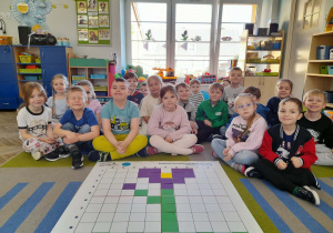 Dzieci siedzą na dywanie, a przed nimi leży mata do kodowania, na której znajduje się ułożony przez dzieci krokus według podanego kodu.