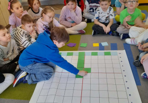Staś siedzi obok maty i dokłada zielony kartonik według kodu. Koleżanki i koledzy obserwują działanie chłopca.