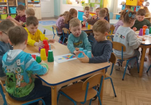 Dzieci siedzą przy stołach i układają wieże według podanych wzorów.